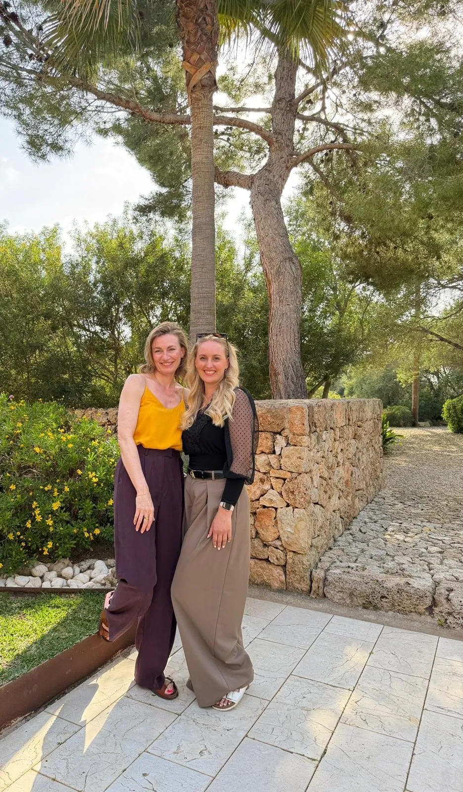 Katrin und Swetlana stehen lächelnd im Garten der Finca auf Mallorca, hinter ihnen Pinien und eine Natursteinmauer